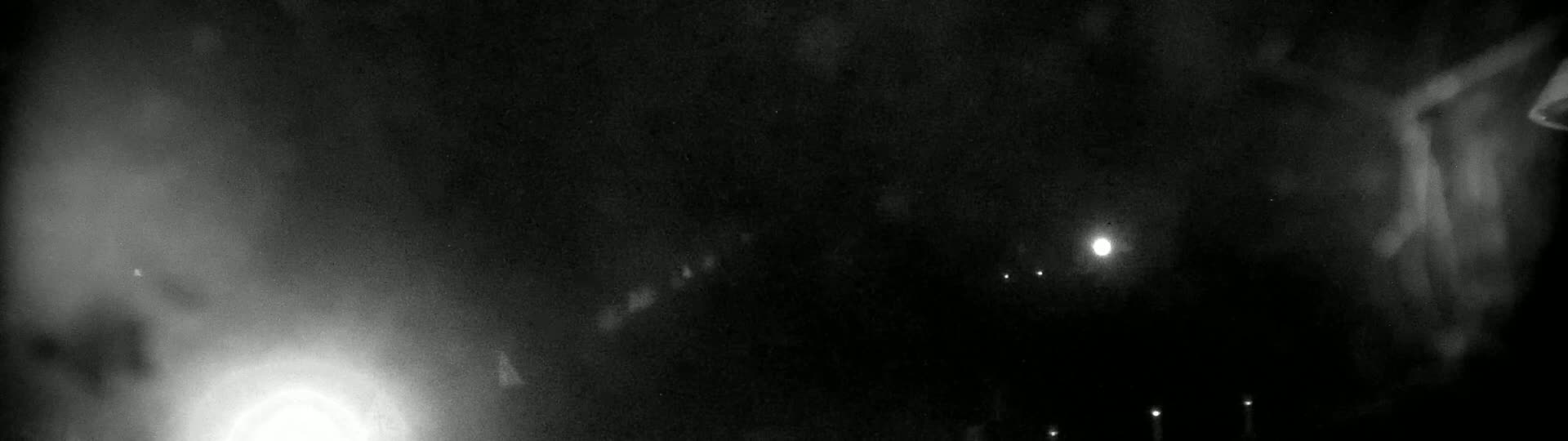 A nighttime, blurry image shows a bright light in the lower left corner and several fainter lights in the distance, possibly streetlights, with a hazy or foggy atmosphere.