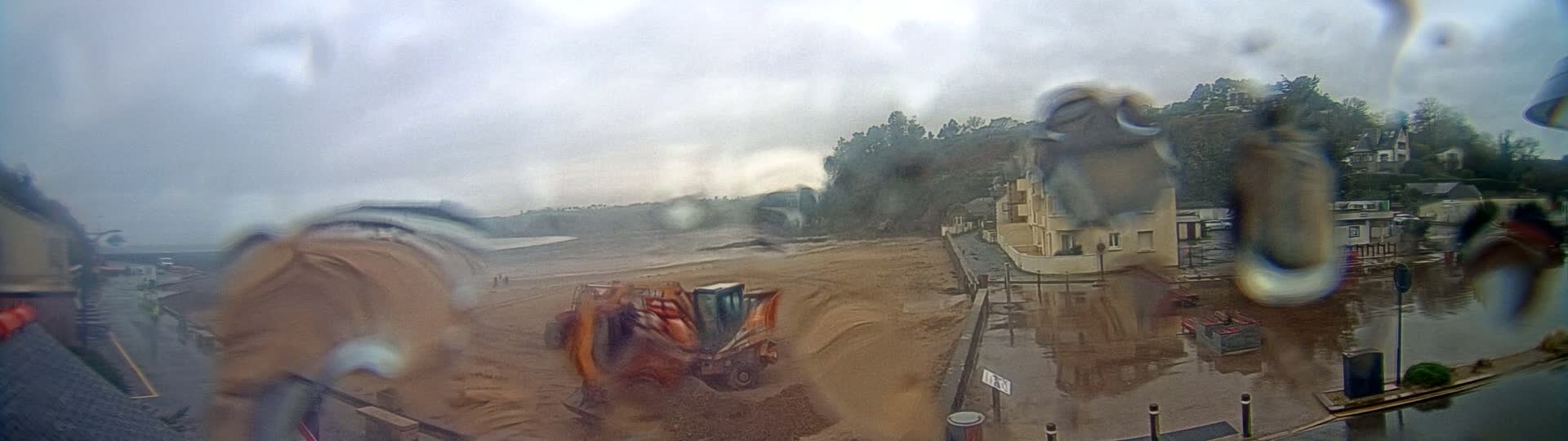 A view through rain-covered glass shows an orange piece of construction equipment working on a beach, with buildings and trees visible in the background under an overcast sky.