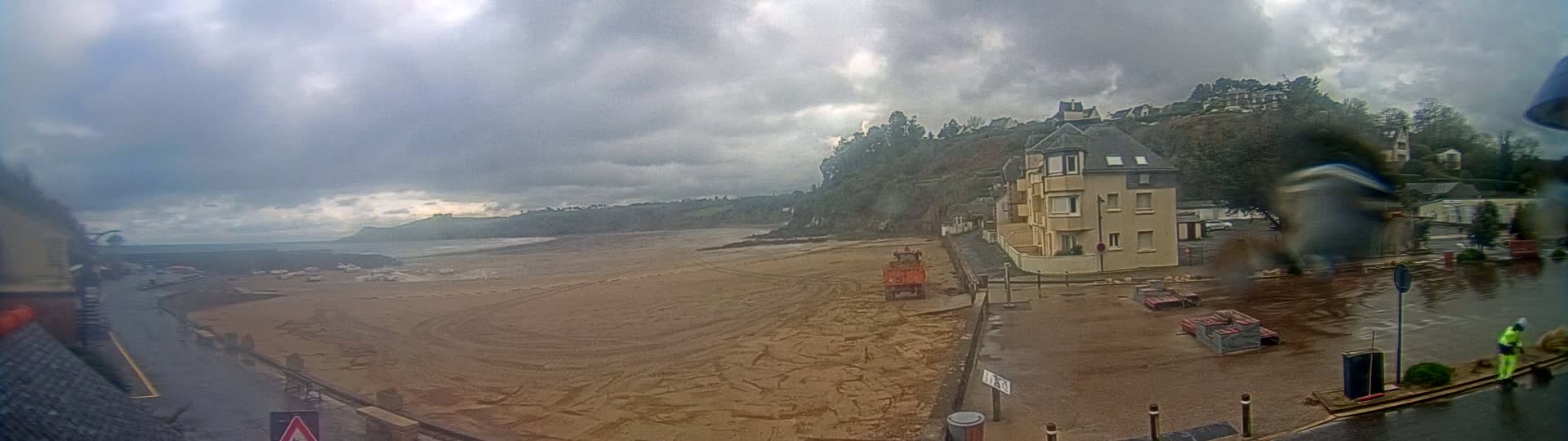 A sandy beach stretches out under a cloudy sky, with a three-story building and construction equipment visible near the water's edge.