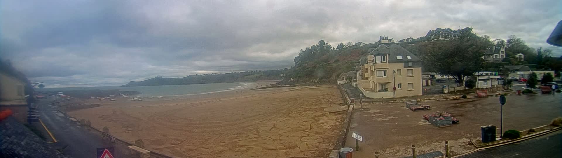A sandy beach stretches out to a calm ocean under a cloudy sky, with buildings and trees on a hillside overlooking the shore.
