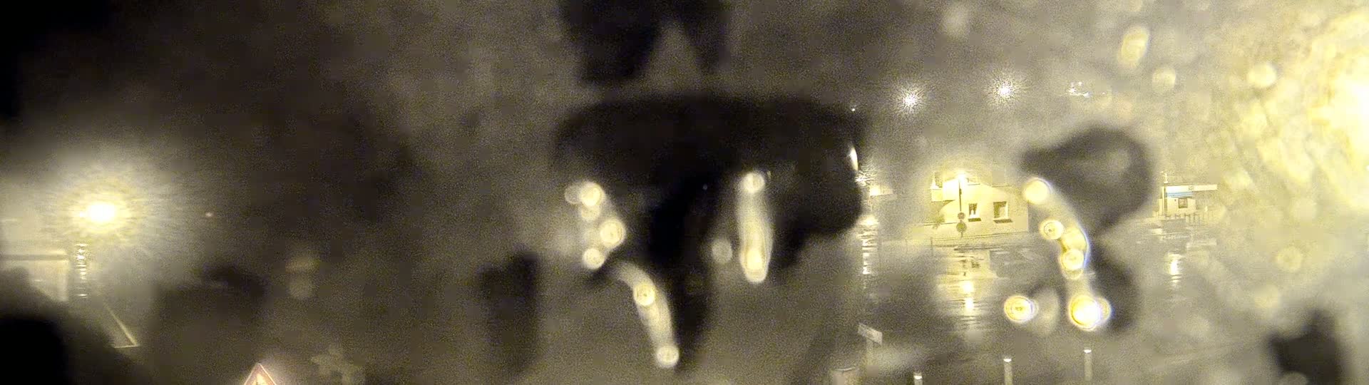 A blurry nighttime view of a street scene seen through a rain-covered window, showing indistinct buildings and lights.