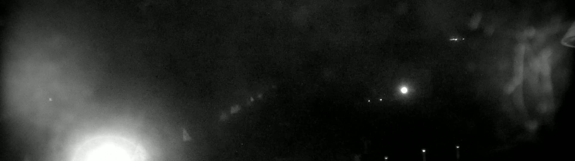 A nighttime, hazy or foggy scene shows various lights in the distance, possibly streetlights or headlights, with a brighter light source on the lower left.