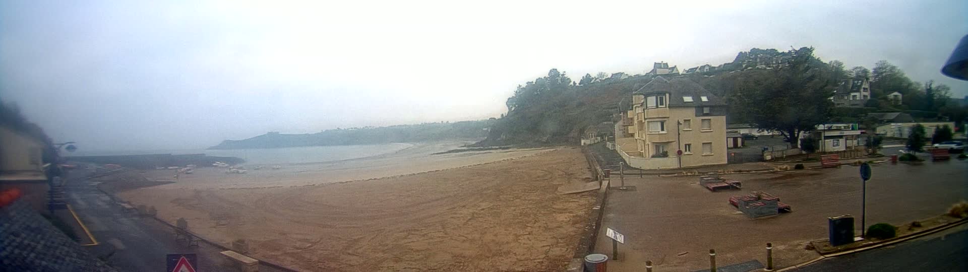 A sandy beach stretches out to a calm, foggy ocean, with a multi-story building and other structures situated on a hill overlooking the shore.