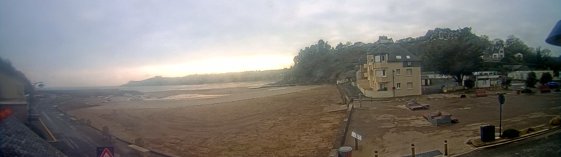 A sandy beach stretches out to a calm body of water under an overcast sky, with buildings and trees visible on a hillside in the background.