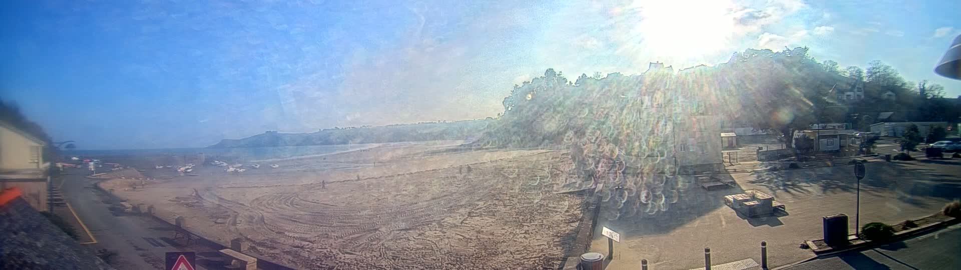 A sunny day at a beach shows a sandy shore with tire tracks, a few people, and boats pulled up on the sand; beyond the beach are buildings and trees on a hillside.