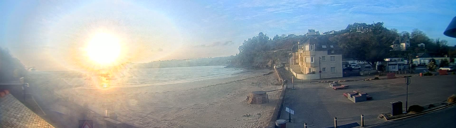 A sandy beach with gentle waves meets a paved area with buildings and park benches, all bathed in bright sunlight.