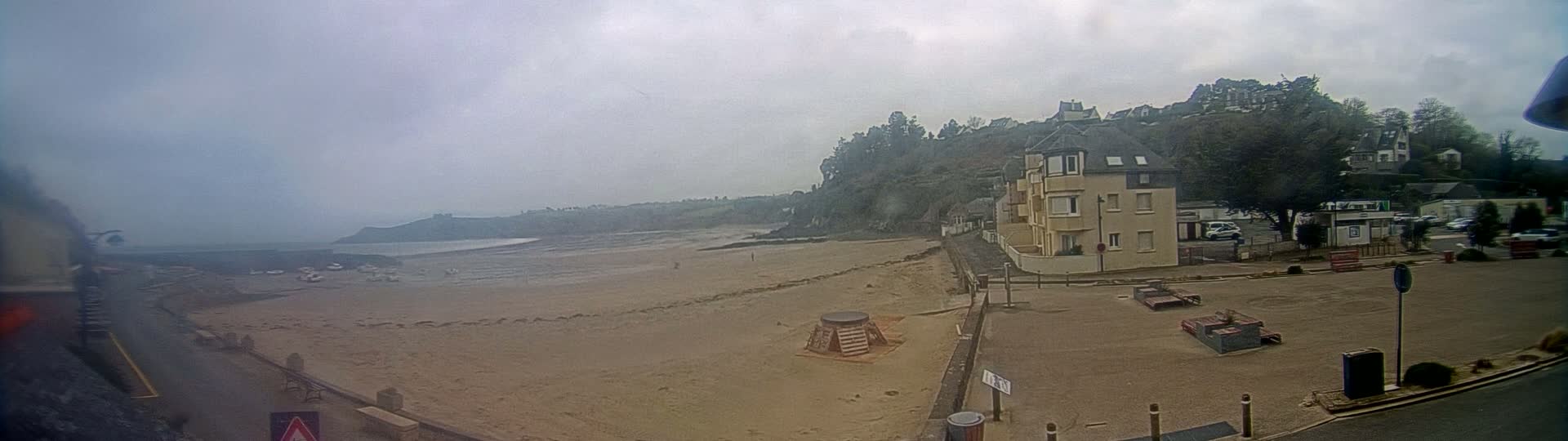 A sandy beach stretches out to a calm ocean under an overcast sky, with buildings and trees rising on a hill in the background.
