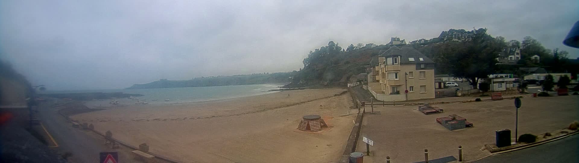 A sandy beach stretches along a calm, gray ocean under an overcast sky, with a multi-story building and other structures visible on a nearby hill.