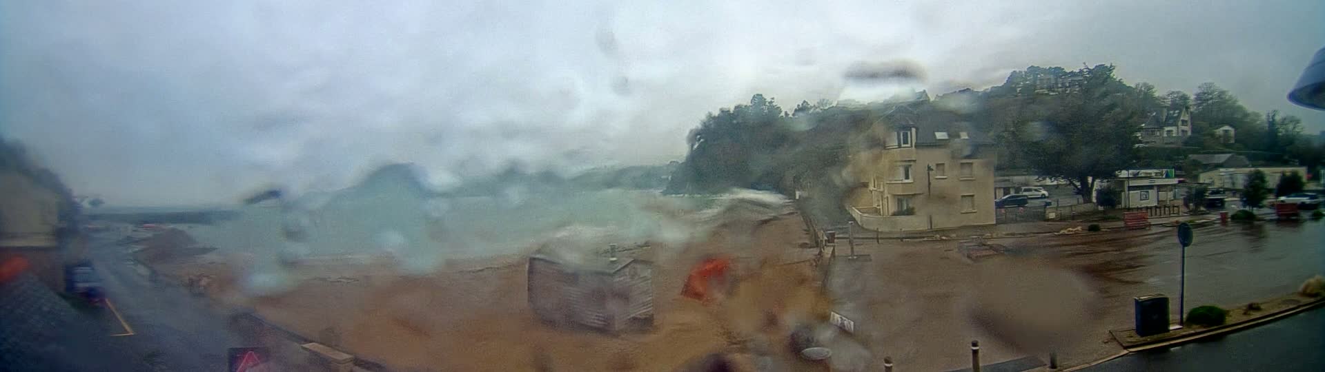 A rain-blurred view shows a coastal scene with a beach, rough waves, a small building, and a multi-story building, all viewed from a window.