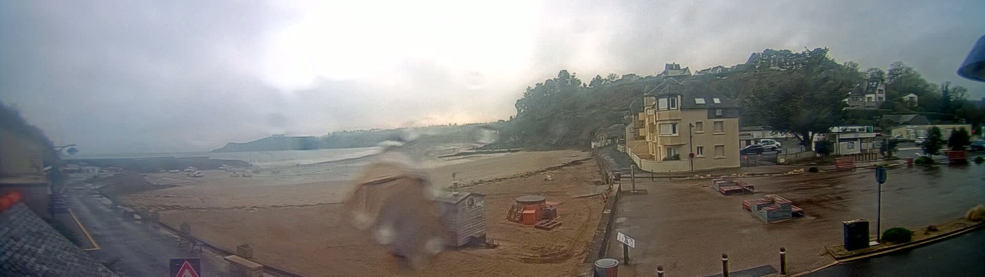 A rain-swept beach scene shows a sandy shore with construction equipment, a small building, and a multi-story building overlooking the water, all under an overcast sky.