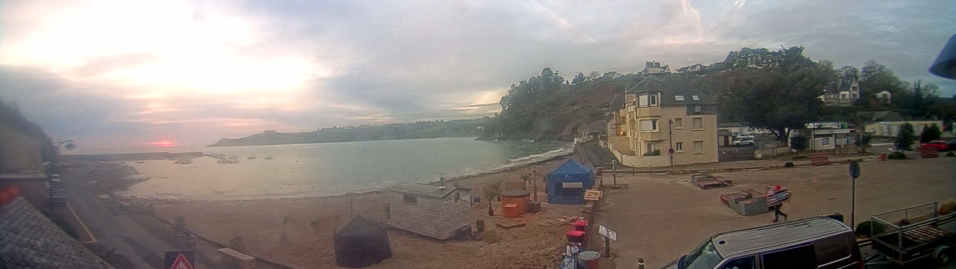 A beach scene at sunset shows calm water, a sandy shore with small buildings and a van, under overcast skies.