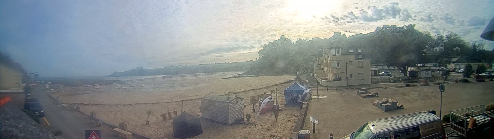 A mostly sunny day reveals a sandy beach at low tide, several small buildings, and a parking area near a hillside with houses and trees.