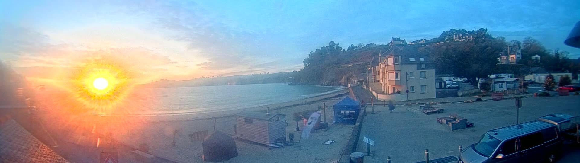 A bright, hazy sunrise over a calm bay with a sandy beach, beach huts, and buildings on a cliffside.