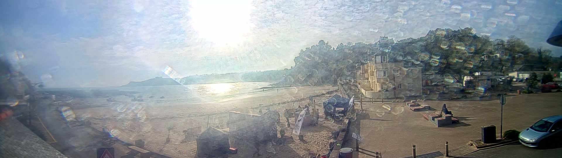 A sandy beach with calm water is visible under a bright, sunny sky, with buildings and people visible in the foreground.