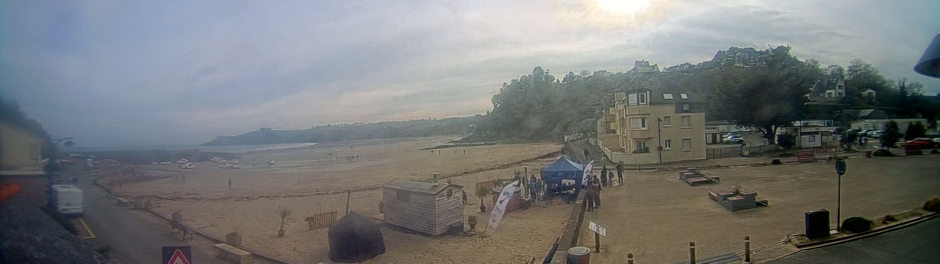 A mostly sunny, hazy day overlooks a sandy beach with a few people and some small structures, near a shoreline with buildings and trees on a hill in the background.