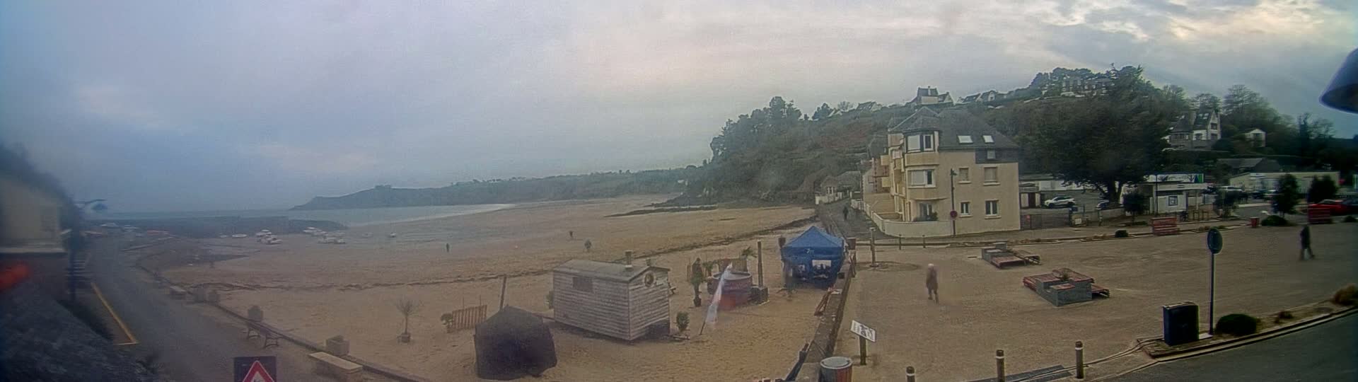 A sandy beach stretches out to a calm ocean under an overcast sky, with a few buildings and people visible along the shore.