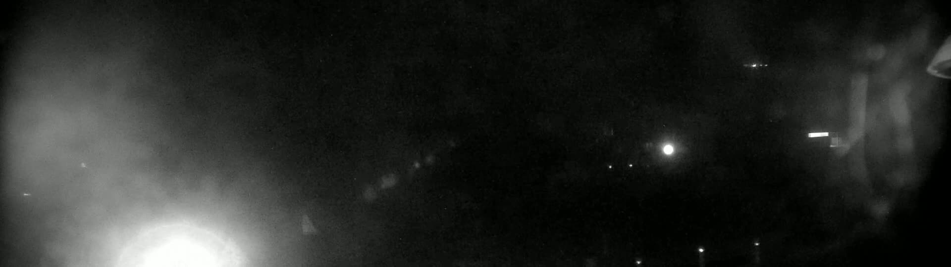 A blurry, nighttime image shows a dimly lit area with several faint lights visible in the distance and a bright light source in the lower left corner.