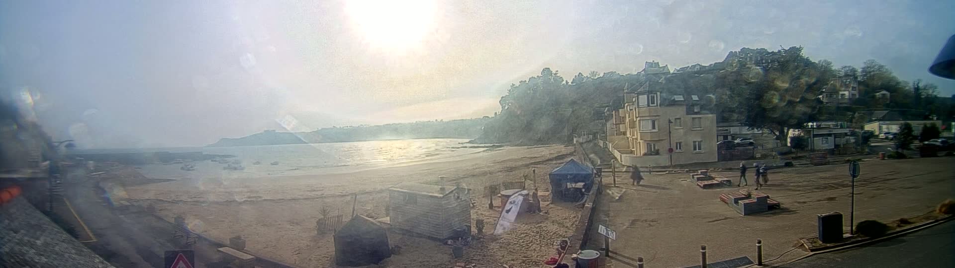 A sandy beach with calm water and a few buildings is seen on a bright, sunny day.