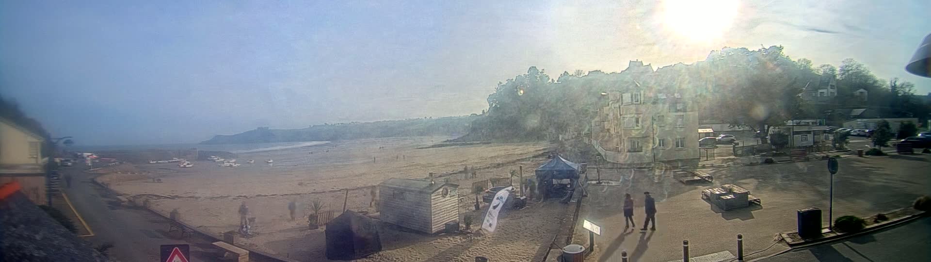 A hazy sunny day overlooks a sandy beach with a few people, small buildings, and parked cars near a seaside town.