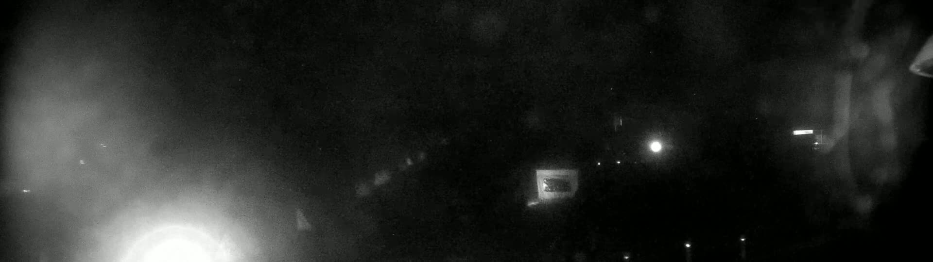 A nighttime, blurry outdoor scene shows a bright light in the lower left corner and several faint lights scattered across a dark background, with a hazy or foggy atmosphere.