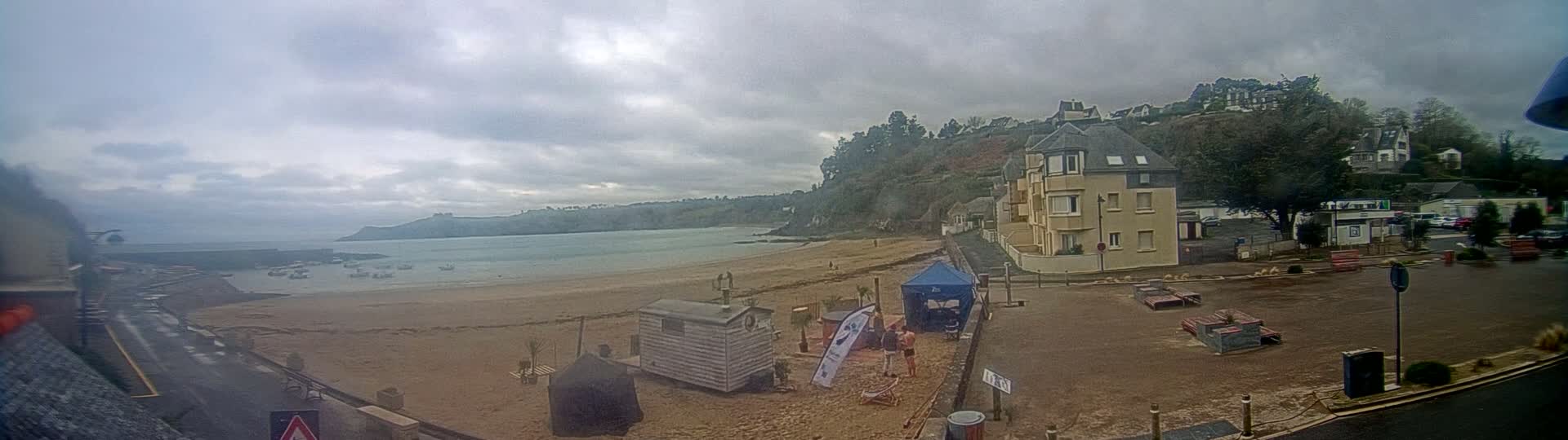 A sandy beach with a few boats in the water and buildings on a hillside overlooking it under an overcast sky.