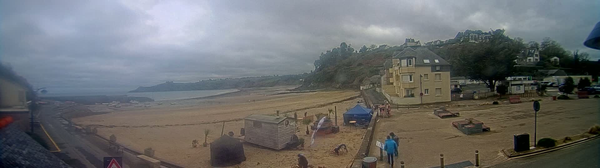 A sandy beach stretches out to a calm ocean under an overcast sky, with buildings and a few people visible along the shoreline.