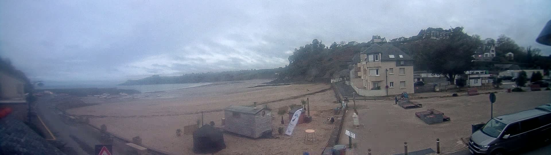 A sandy beach stretches out to a calm ocean under an overcast sky, with buildings and trees lining the shore.