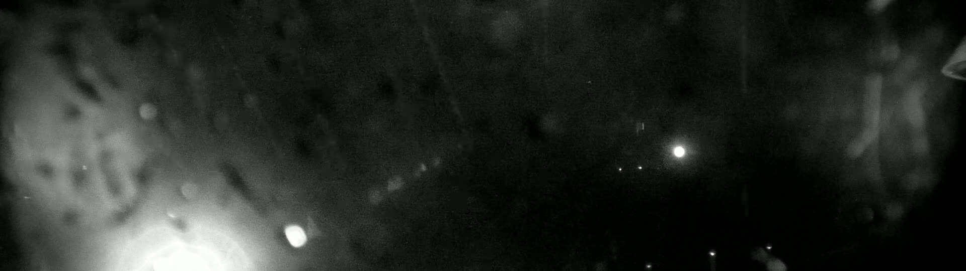 A blurry, dark, nighttime scene shows a few bright lights and what appears to be rain or snow falling.