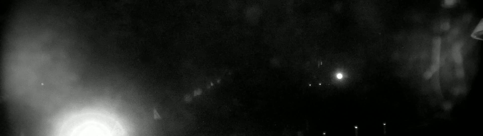 A grainy, dark nighttime image shows blurry lights in the distance, possibly from streetlights or other distant sources, with a brighter light source possibly a headlight or streetlight much closer to the viewer.