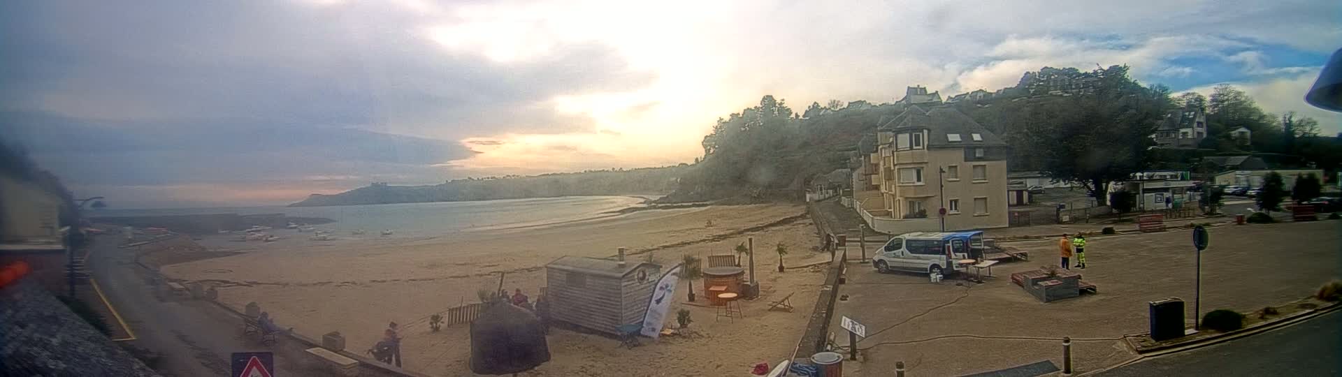 A sandy beach with calm water stretches out under a cloudy sky, with buildings and people visible on the shore.