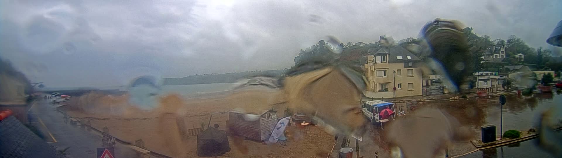 A rain-streaked view shows a beach with a few structures and a building overlooking it, all under a cloudy sky.