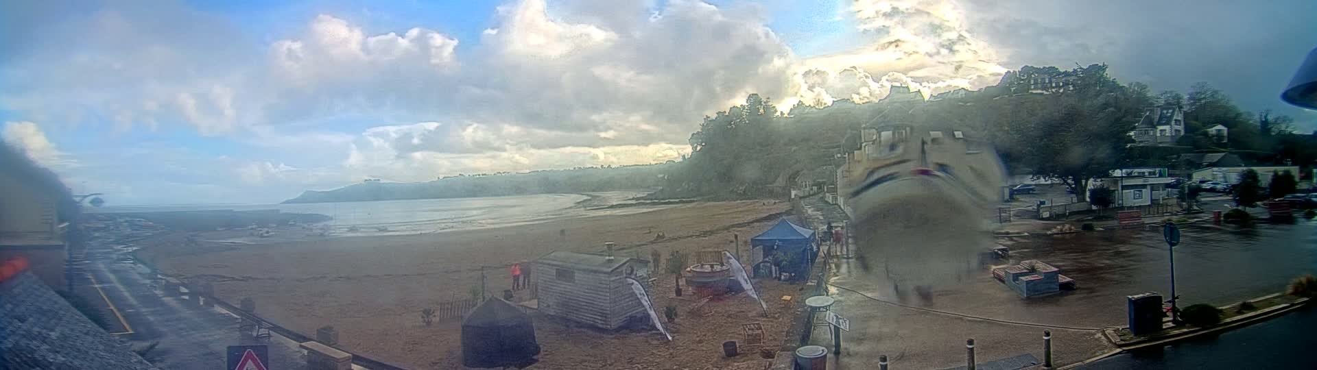 A sandy beach with a few people and small structures is visible, backed by a treeline and buildings on a slightly overcast day with some rain.