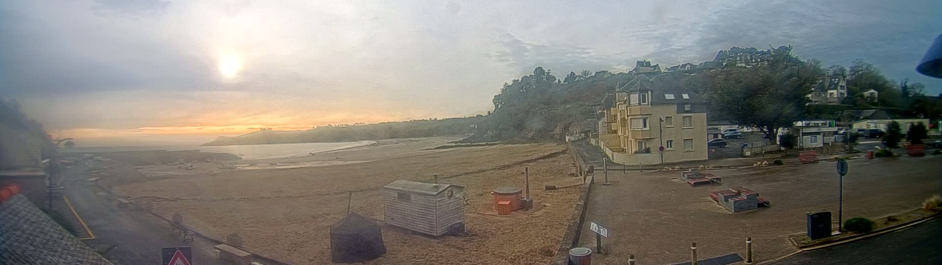A sandy beach stretches out to a calm ocean under an overcast sky, with a building and some parked cars visible in the foreground.