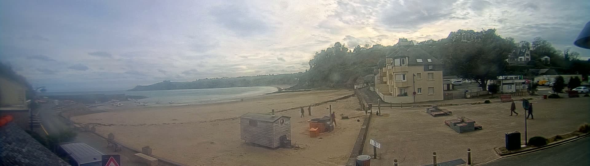A sandy beach stretches along a calm ocean under a cloudy sky, with a few people walking near a building and small structures on the shore.