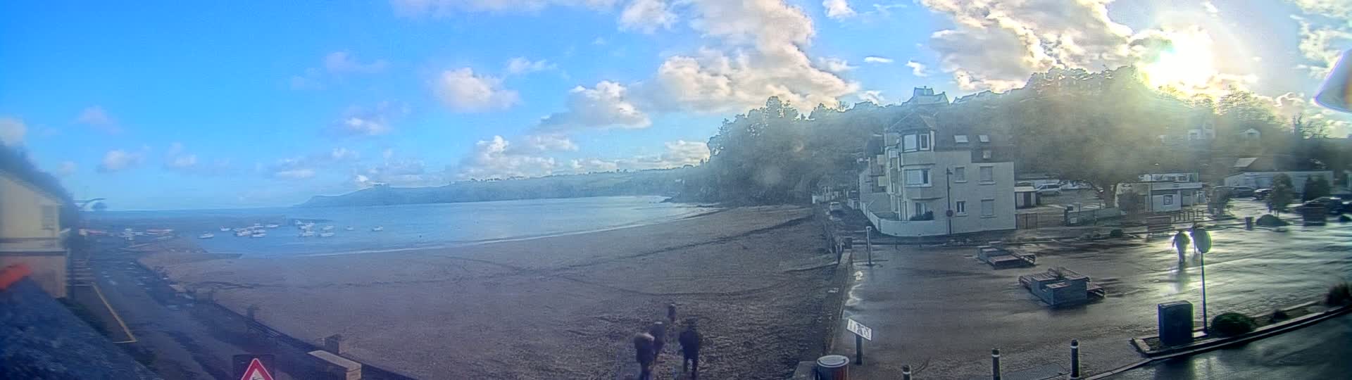 A mostly sunny day shows a sandy beach with calm water, boats in the bay, and buildings on a hillside overlooking the coast.