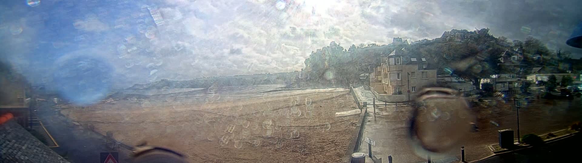 A beach with buildings and trees on a cloudy day is viewed through a rain-splattered window.