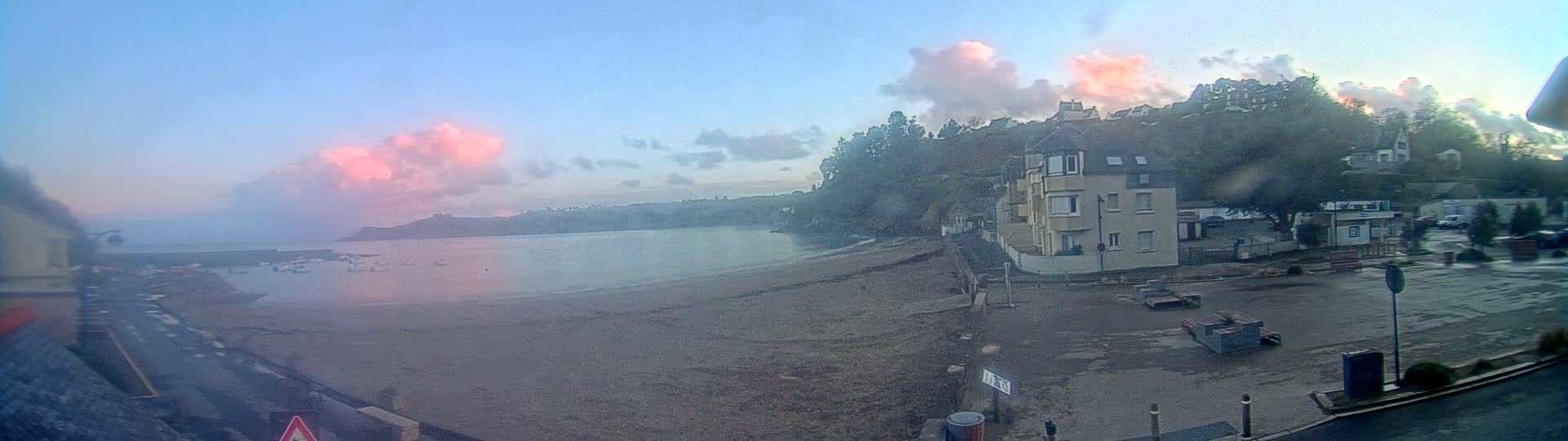 A seaside town with pink-hued clouds in the sky, a calm body of water, a sandy beach, and buildings situated on a hillside, viewed from a slightly elevated perspective in wet conditions.