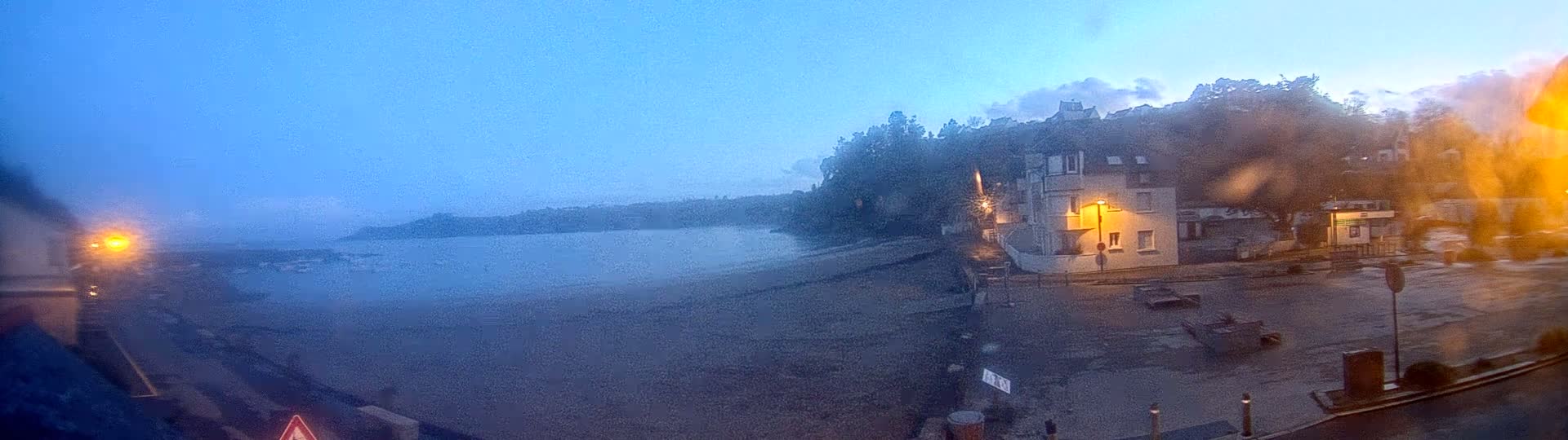 A dimly lit beach at dawn or dusk is seen from a high vantage point, with a building and trees visible on a hill overlooking the water.