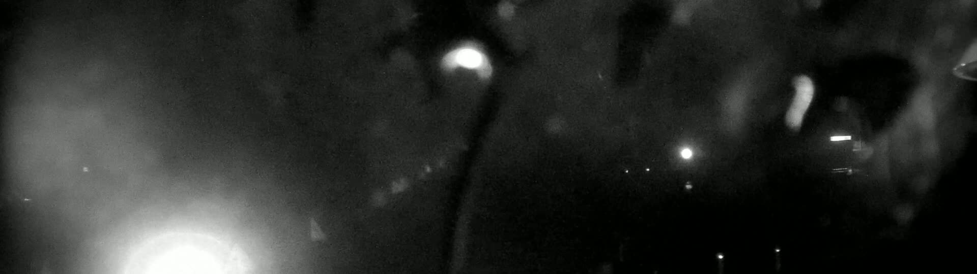 A blurry, nighttime outdoor scene shows rain falling, with indistinct lights visible in the distance.