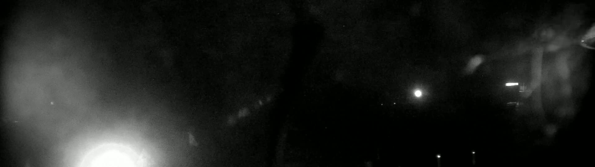 A nighttime, blurry outdoor scene shows a bright light on the lower left, several indistinct lights in the distance, and what appears to be rain or snow falling.