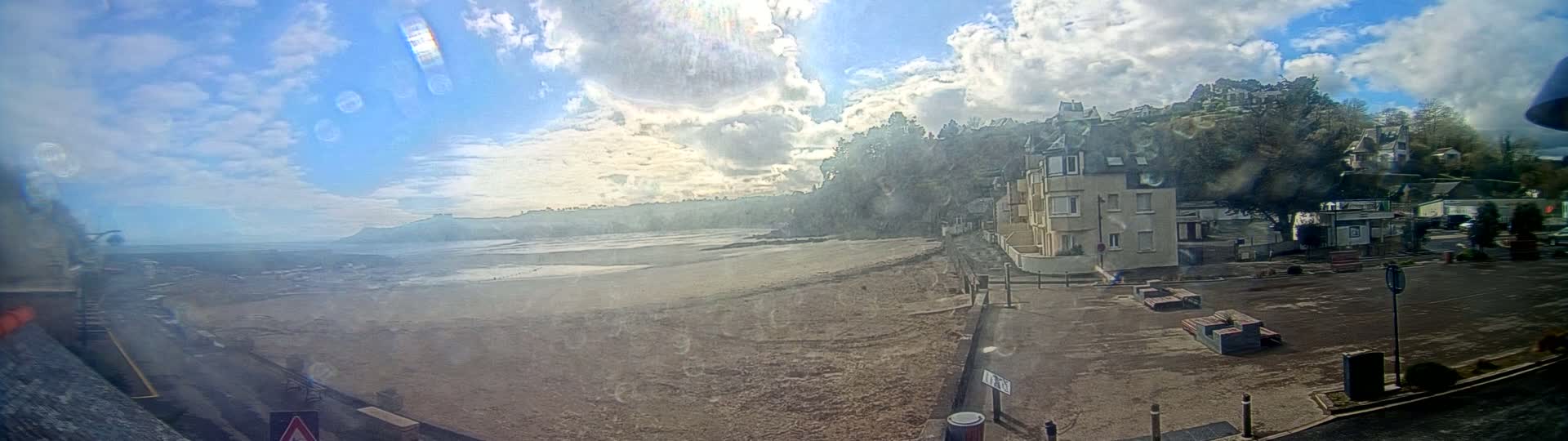 A mostly sunny day with partly cloudy skies shows an expansive sandy beach, a building overlooking the beach, and a parking lot, all viewed through a rain-streaked window.