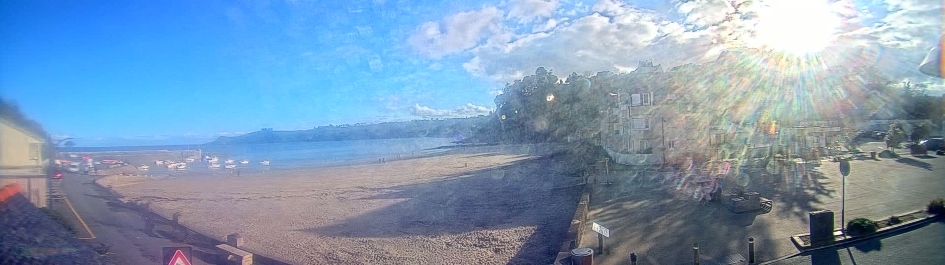 A sandy beach with several small boats is visible from a coastal road, with buildings and trees in the background under a bright, sunny sky with some clouds.
