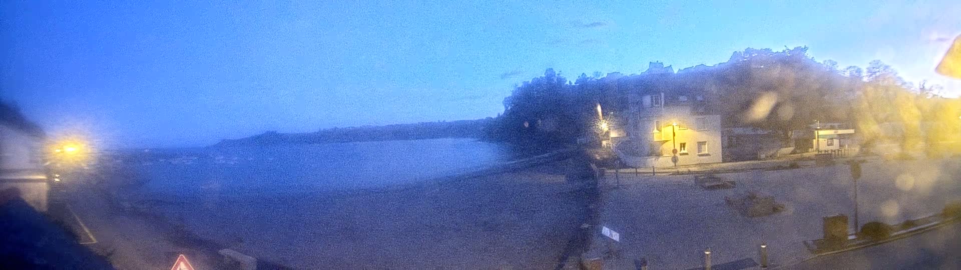 A blurry, nighttime view of a waterfront town with a building lit by a streetlamp, under a clear, twilight sky.