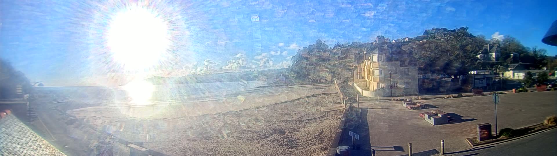 A sunny day shows a sandy beach next to a paved area with buildings and trees on a hillside in the background.
