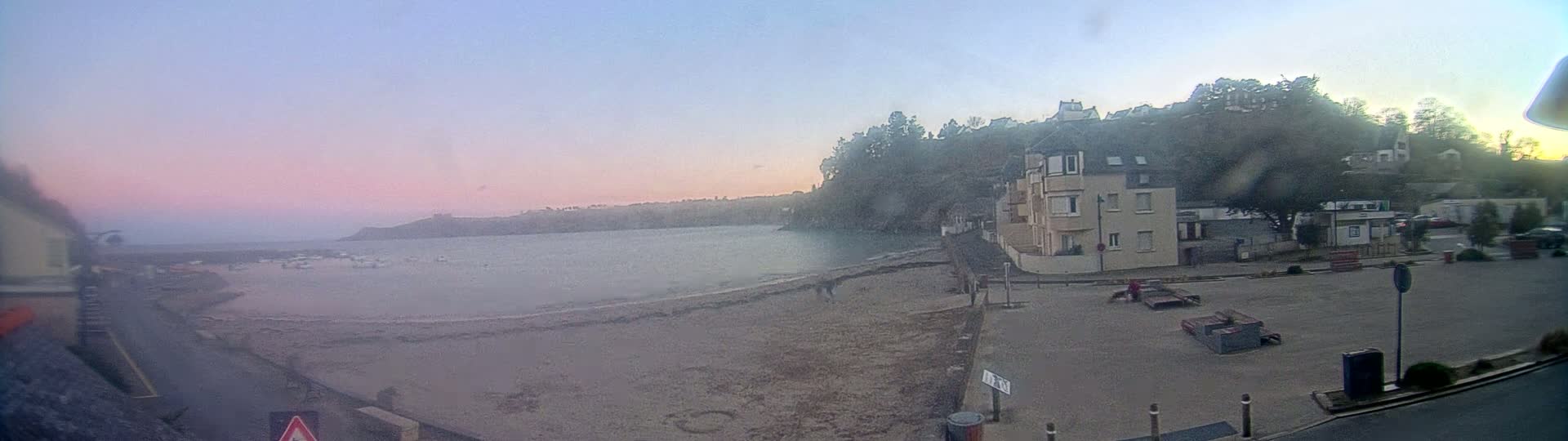 A calm bay with several boats, a sandy beach, and a residential building overlooking the water at sunset.