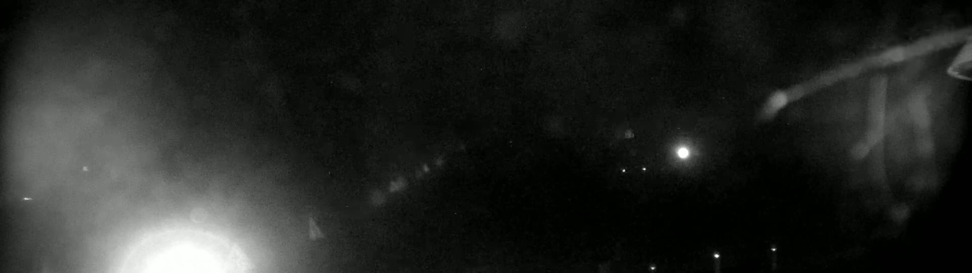 A nighttime, blurry, outdoor scene shows a bright light in the lower left corner and several fainter lights in the distance, with a dark, possibly rainy or foggy, sky.