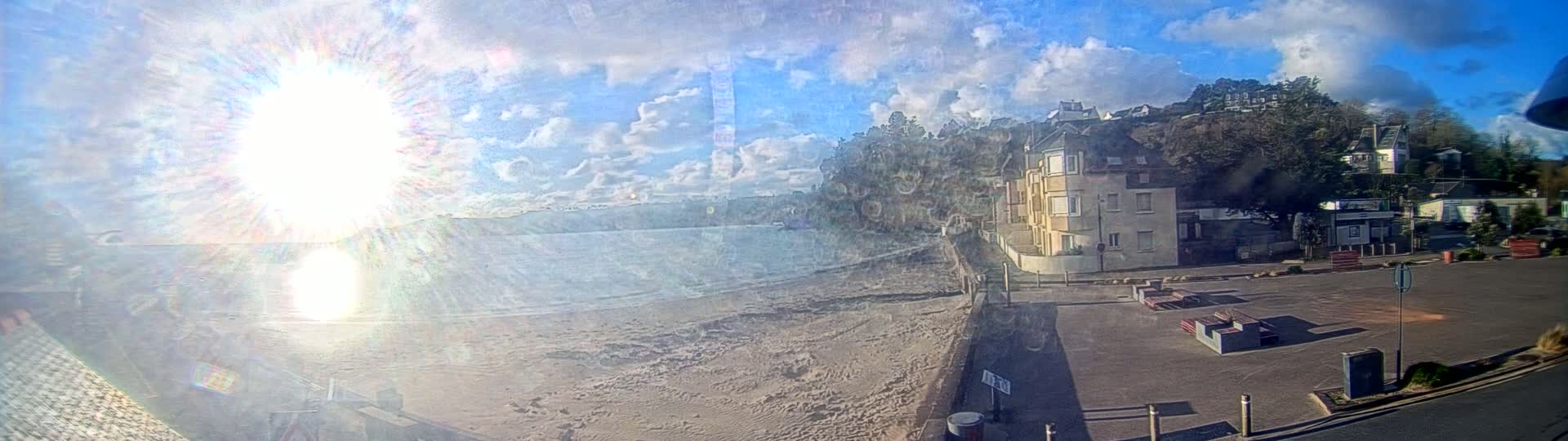 A sandy beach stretches out to a calm body of water under a bright, sunny sky with scattered clouds, next to a parking lot and buildings on a hillside.