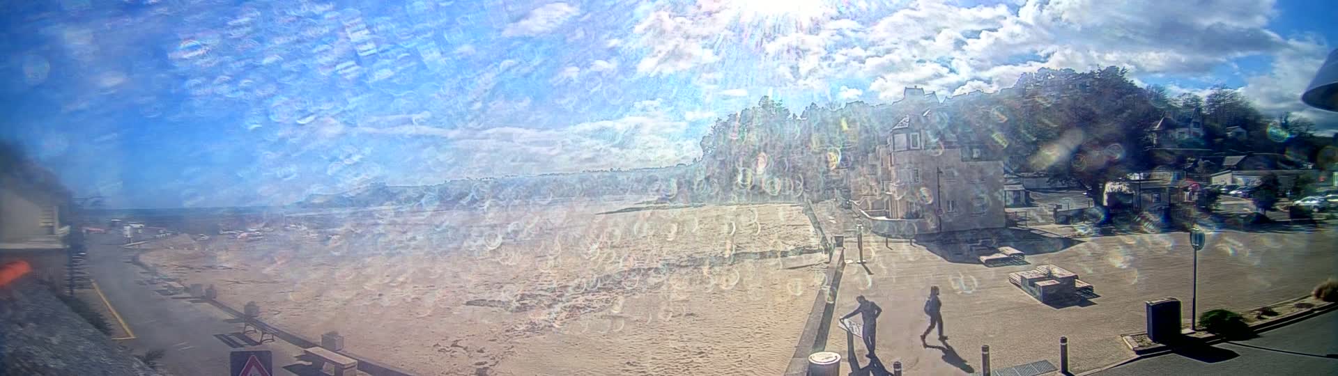 A sandy beach stretches out beneath a bright, sunny sky with scattered clouds, alongside a paved area with buildings and a few people walking.