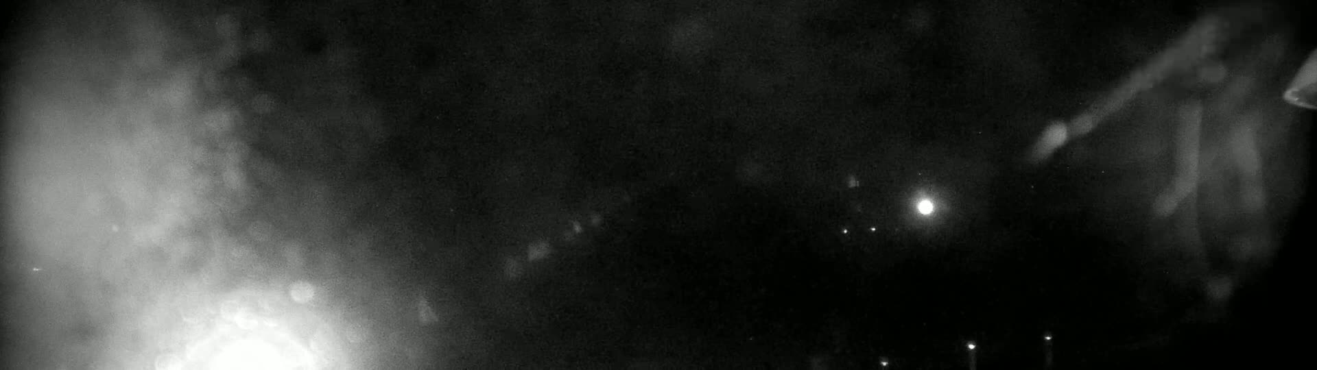 A blurry, nighttime image shows bright lights in the distance and indistinct shapes in the foreground under a dark, possibly hazy or snowy sky.