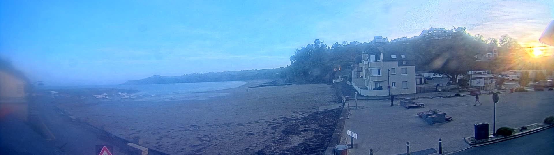 A sandy beach stretches to a calm body of water under a clear, bright blue sky with a setting sun.